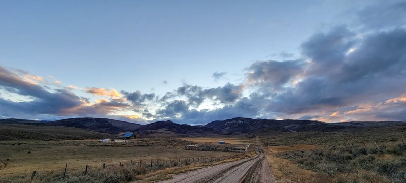 Dirt ranch road leading to a blue-roofed barn under sunset clouds